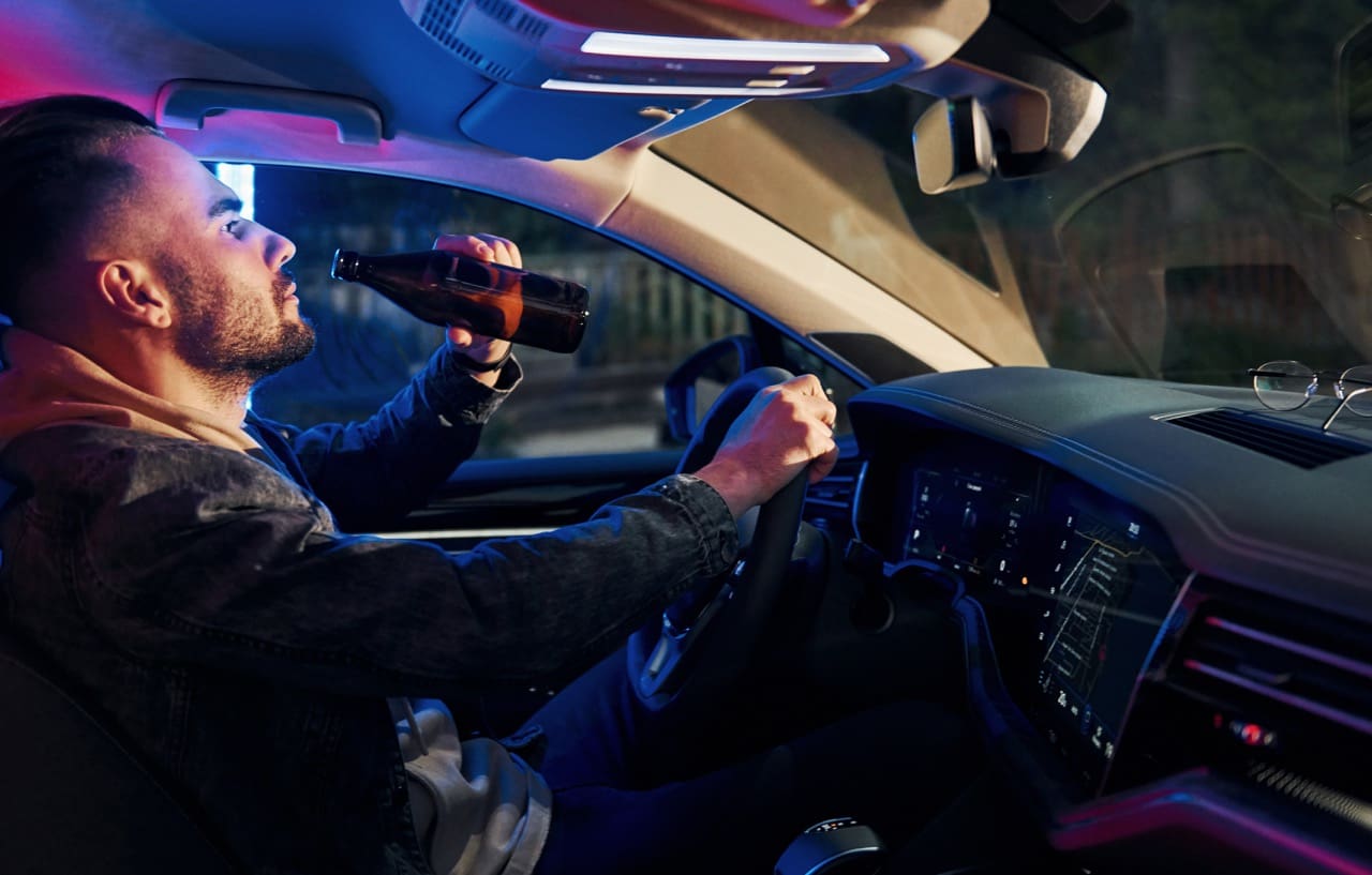 Side view of a man drinking and driving. Police lights shine in the background