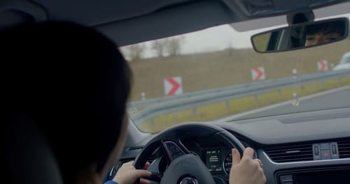 View from the passenger seat of a car showing the driver holding the steering wheel, looking ahead at a road with curve warning signs.