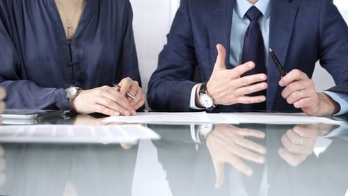 Two people in business attire sit at a glass table, discussing documents; one gestures with hands while the other holds a pen.