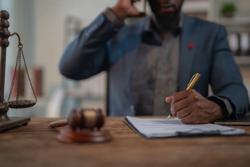 A person in a suit talks on the phone while writing on a document at a desk with a gavel and a scale of justice.