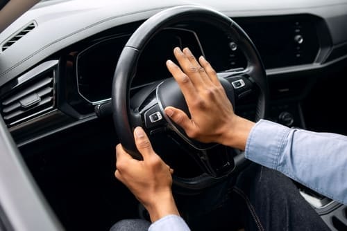 Person pressing the horn on a car steering wheel with one hand while holding the wheel with the other hand, inside a modern vehicle.