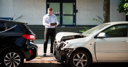 A man stands on the sidewalk using a tablet while inspecting the scene of a collision between a black car and a white car with front-end damage.