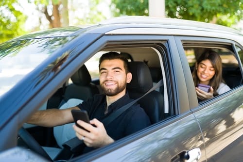 A man in the driver's seat and a woman in the back seat of a car are both using their smartphones. The car is parked near trees on a sunny day.