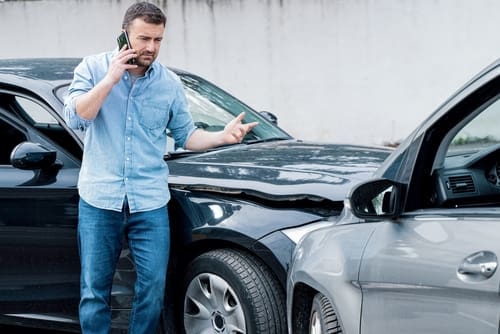 A man stands next to two cars involved in an accident, talking on his phone and gesturing toward the damaged vehicles.
