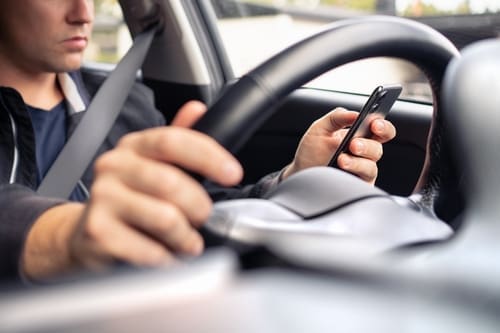 A person driving a car with one hand on the steering wheel and the other hand holding a smartphone.