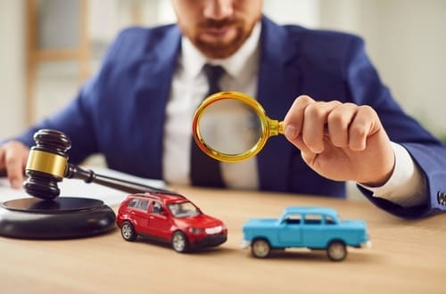 A person in a suit examines two toy cars with a magnifying glass next to a judge's gavel on a desk.