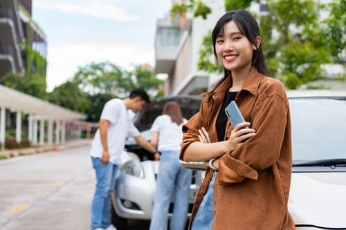 A woman stands smiling in the foreground holding a phone, while two people check under the hood of a car in the background.