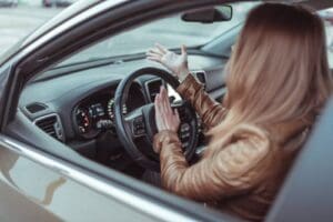 A woman sits in the driver’s seat of a car with her hands raised, appearing frustrated or confused, while looking out the window.