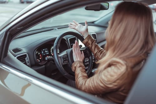 A woman sits in the driver’s seat of a car with her hands raised, appearing frustrated or confused, while looking out the window.