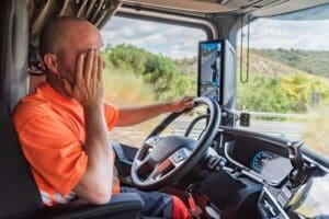 A truck driver in an orange shirt sits in the driver's seat, holding the steering wheel with one hand and rubbing his face with the other.