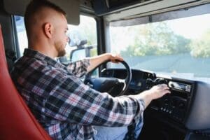 A man wearing a plaid shirt is seated in a truck, driving with one hand on the wheel and the other hand adjusting the dashboard controls.