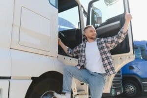 A man standing on a truck step with one hand on the door and giving a thumbs up, smiling and looking away.