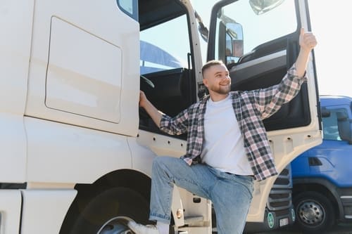 A man standing on a truck step with one hand on the door and giving a thumbs up, smiling and looking away.