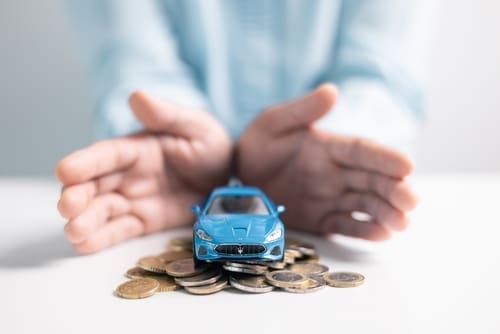 A person’s hands protectively encircle a blue toy car placed on top of a pile of coins on a white surface.