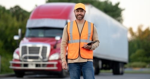 Man wearing an orange safety vest and yellow cap stands in front of a red and white semi-truck, holding a tablet.
