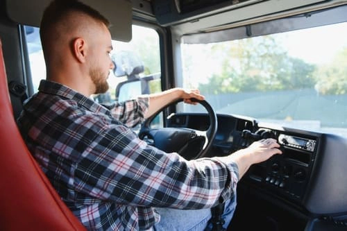 Man in a plaid shirt sits in the driver's seat of a truck, holding the steering wheel with one hand and adjusting controls on the dashboard with the other.