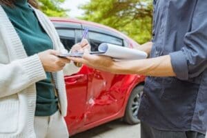 Charlotte Hit and Run Accident Lawyer - Payne Law Firm Two people stand near a red car with a dented door; one person is signing a document on a clipboard held by the other.