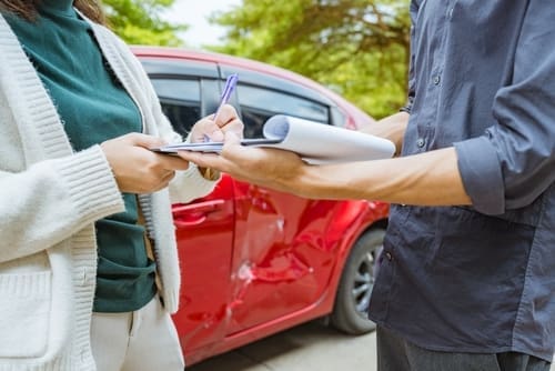 Two people stand near a red car with a dented door; one person is signing a document on a clipboard held by the other.