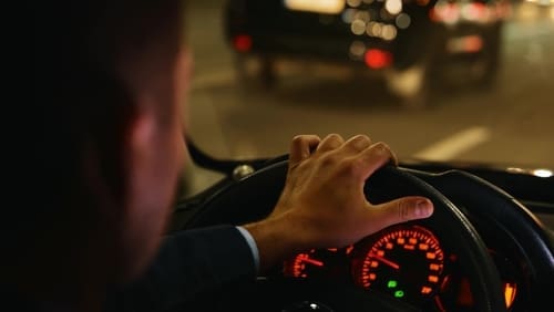 A person driving a car at night, with one hand on the steering wheel and the dashboard illuminated, following another vehicle on the road.