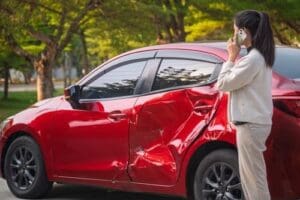 Charlotte Hit and Run Accident Lawyer - Payne Law Firm A woman stands next to a red car with a large dent on the side, talking on her phone.