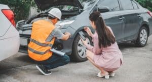 Charlotte Hit and Run Accident Lawyer - Payne Law Firm A man in a safety vest inspects the bumper of a gray car with its hood open while a woman kneels nearby, taking a photo with her phone.