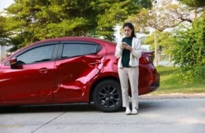Charlotte Hit and Run Accident Lawyer - Payne Law Firm A woman stands next to a red car with a large dent on the rear door, looking at her phone in an outdoor parking area.