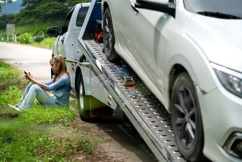 A woman sits on the grass beside a road, using her phone, while a white car is being loaded onto a tow truck.