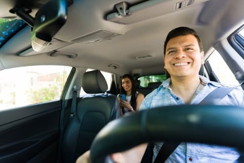 A man is driving a car while a woman sits in the back seat, looking at her phone. Both appear to be passengers in a rideshare vehicle during the day.