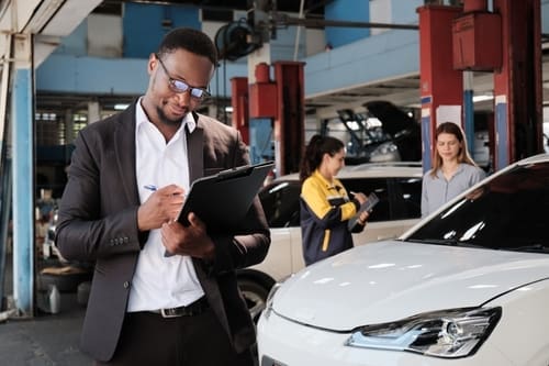 A man in a suit writes on a clipboard in an auto repair shop, with workers and cars visible in the background.