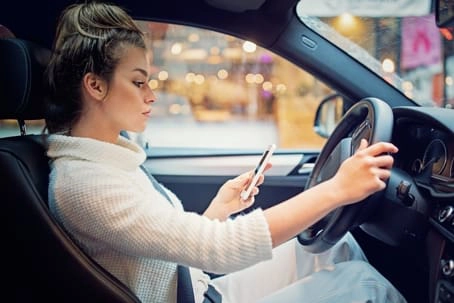 Woman sitting in the driver’s seat of a car, looking at her phone while holding the steering wheel with one hand.