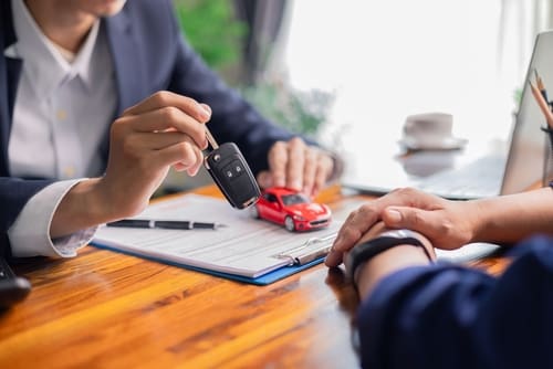 A person hands over a car key to another person across a desk with paperwork and a small red car model.