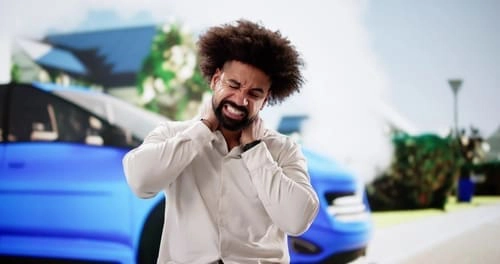A man in a white shirt holds his neck in pain while standing in front of a blue car and suburban background.