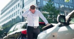 A man stands between two cars after a rear-end collision on a city street, looking down with one hand on the back of his neck.
