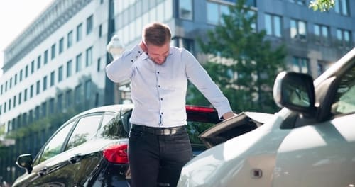 A man stands between two cars after a rear-end collision on a city street, looking down with one hand on the back of his neck.