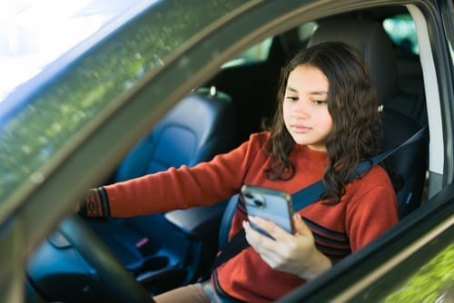 Teen sitting in the driver's seat of a car, wearing a seatbelt, and holding a smartphone while looking at the screen.
