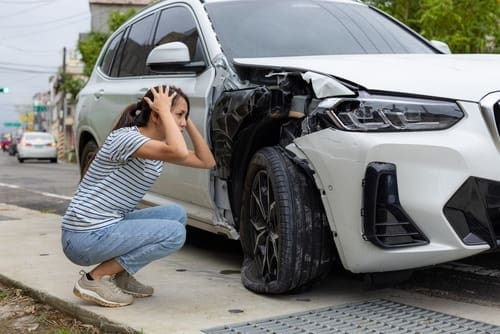 A woman crouches on the sidewalk with her hands on her head next to a white car with significant front-end damage.