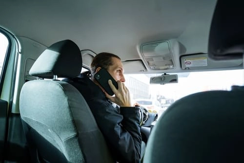 A man is driving a car and talking on a mobile phone, viewed from the back seat during the day.