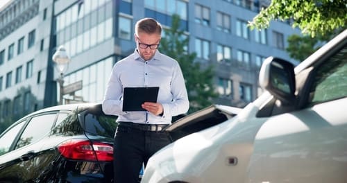 A man in business attire stands between two cars, looking at a tablet, likely documenting or assessing a car accident on a city street.