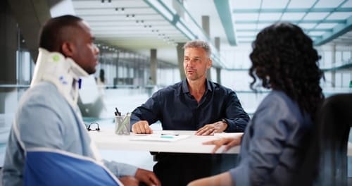 A man with a neck brace and arm sling sits at a desk with a woman, speaking to another man in an office setting.