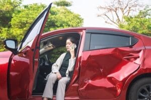 Charlotte Hit and Run Accident Lawyer - Payne Law Firm A woman sits in a red car with a large dented door, talking on a cellphone with the driver’s door open.