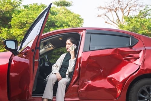 A woman sits in a red car with a large dented door, talking on a cellphone with the driver’s door open.