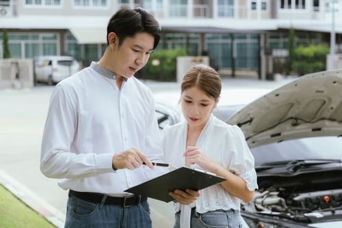 Two people stand by a car with its hood open, reviewing documents on a clipboard, possibly discussing car repairs or an insurance claim.
