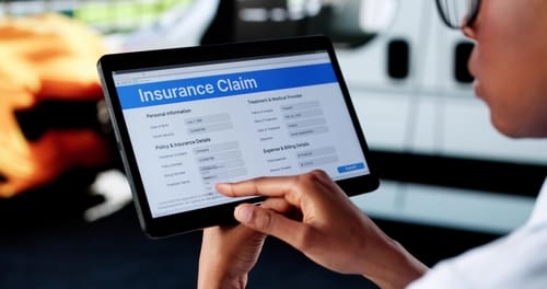 A person holding a tablet fills out an online insurance claim form, with fields for personal information and insurance details visible on the screen.