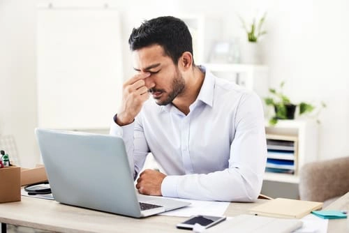 A man sits at a desk with a laptop, pinching the bridge of his nose and appearing stressed or fatigued.