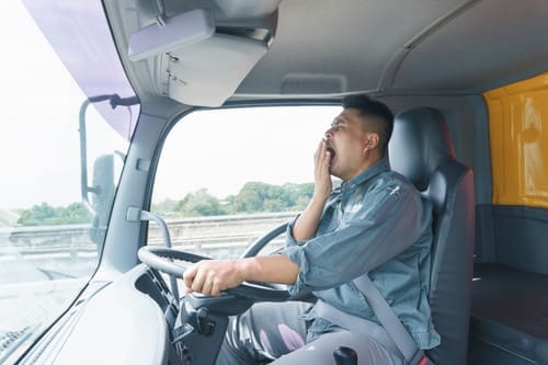 A man sitting in the driver's seat of a truck is yawning and covering his mouth while driving on a road during the day.