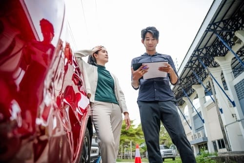 A man holding a clipboard talks to a woman standing next to a red car with visible damage, outdoors near a building.