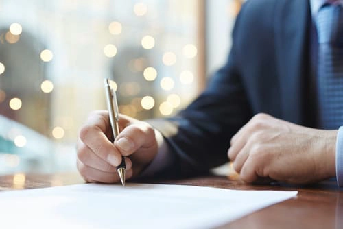 A person in a suit writes with a pen on a sheet of paper at a wooden desk, with blurred lights in the background.