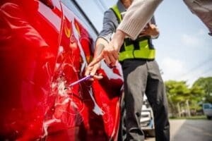 A person points at scratches and damage on the side of a red car while another person in a safety vest takes notes.