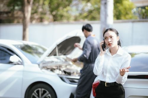 A woman talks on her phone looking frustrated while a man inspects the engine of a white car with its hood up.