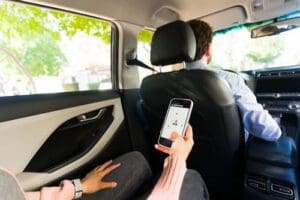 A person in the back seat of a car holds a smartphone displaying a ride-hailing app while a driver sits in the front seat.